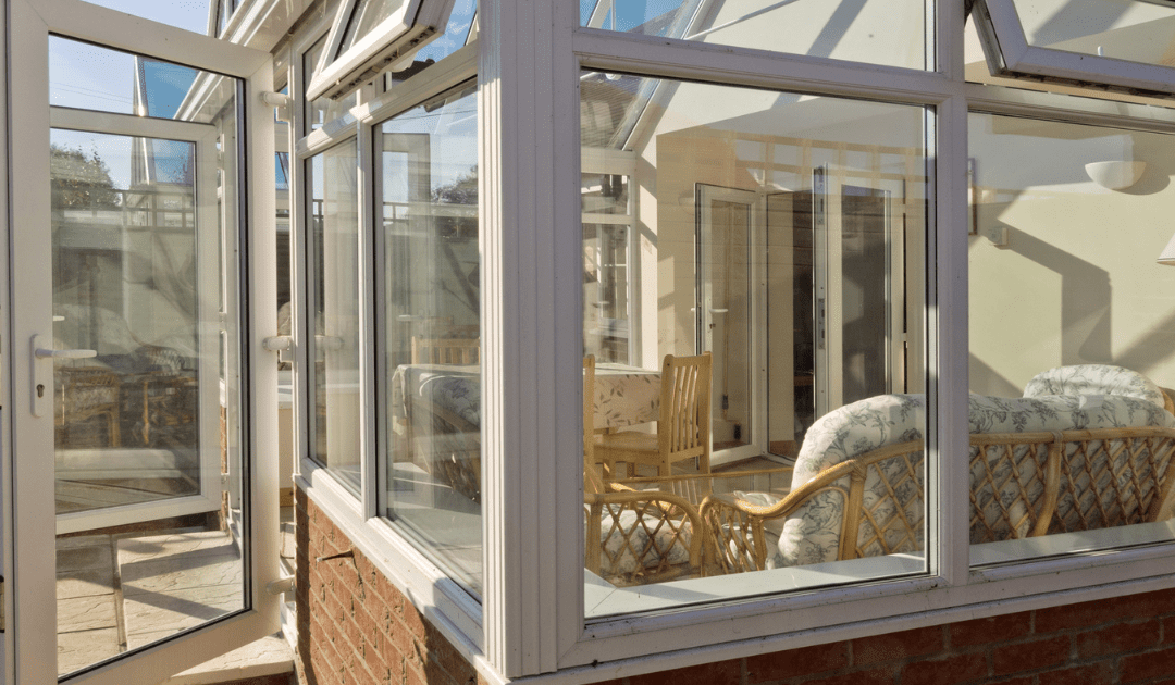 A sunlit conservatory with large glass windows, wicker furniture, and a dining set inside. The open door invites a warm, tranquil ambiance with greenery outside.