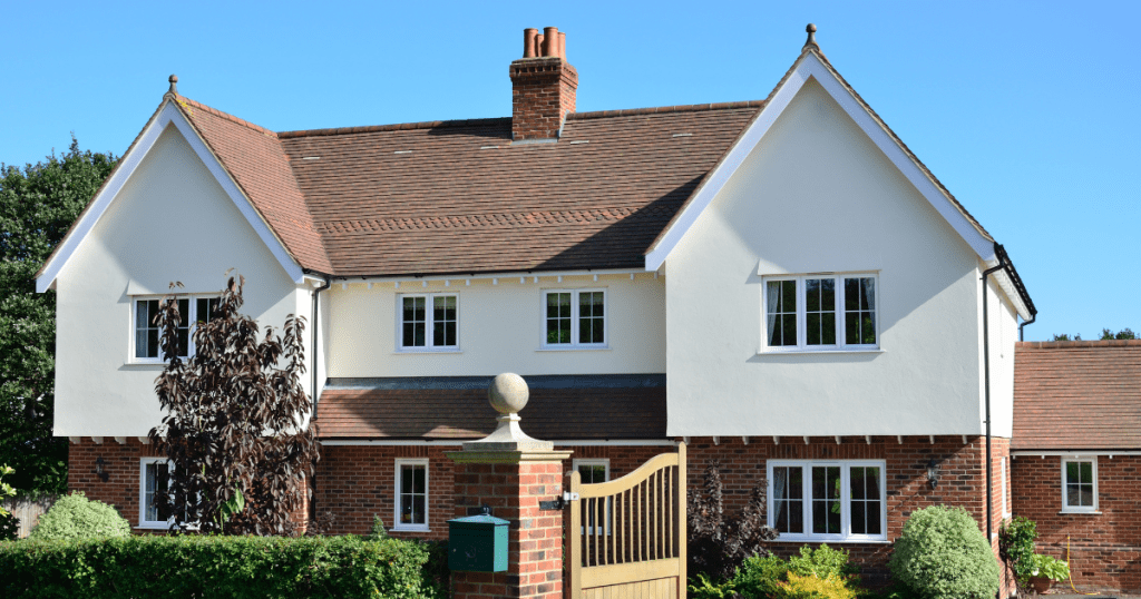 A charming two-story house with white walls and a red-tiled roof. The front yard features shrubs and a small tree, with a wooden gate. Bright, sunny day.